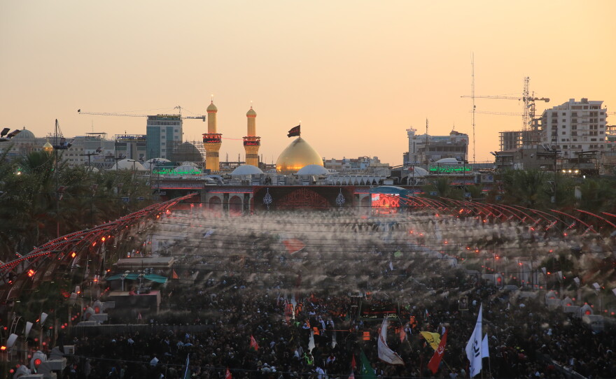 Large crowds of pilgrims fill the area between the two holy shrines in Karbala at sunset, with the illuminated dome and minarets of Imam Hussein's shrine visible in the background during the Arbaeen commemoration.