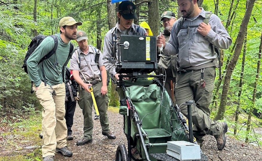 A crew of outdoor industry professionals huddle around a HETAP cart in the rain for a training session at the Delaware Water Gap. A blue umbrella protects the laptop and other technology housed in the rolling device as it's pushed down a tree lined trail. (left to right: Gus Parrish, Jesse Rourke, Matthew Clarke, Christopher Dubas, and Garret Wilson).