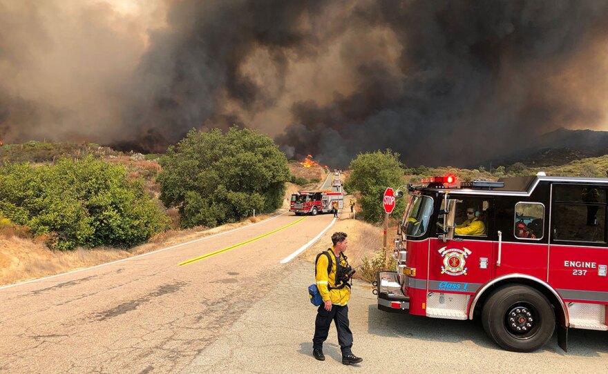 Firefighters block the road as the flames from the Valley Fire burn behind them. The wildfire is located at Japatul Road and Carveacre Road, southeast of Alpine in San Diego County. Sept. 6, 2020.