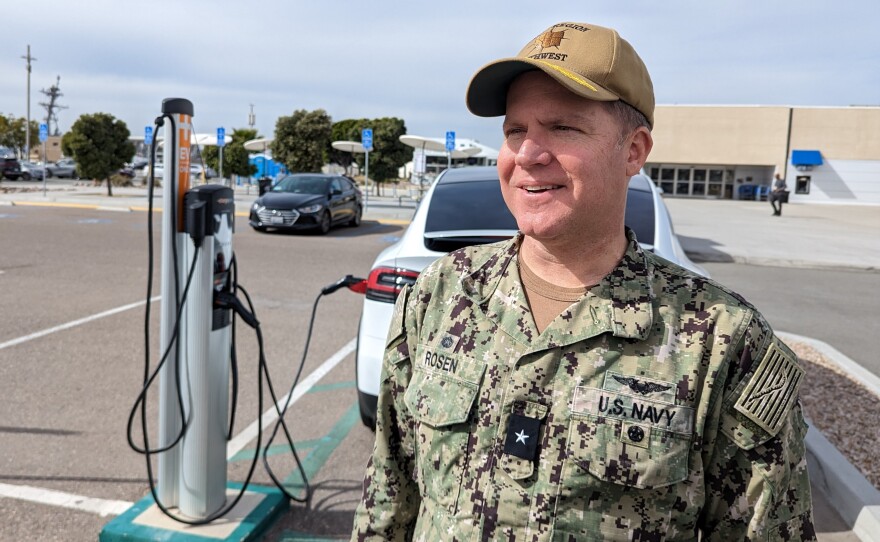 Rear Adm. Brad Rosen, dressed in a green camouflage Navy uniform, stands behind a Tesla charging at a station on Naval Base San Diego on Feb. 17, 2023. 