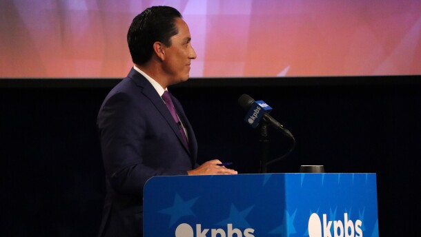 San Diego Mayor Todd Gloria speaks at a podium with the KPBS logo on it. He's wearing a navy blue suit jacket, white shirt and purple tie. 