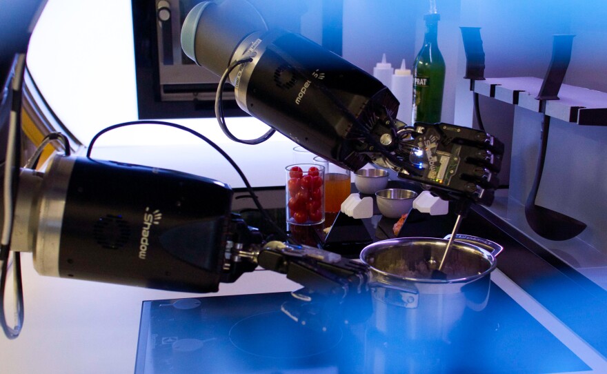 The robotic arms, part of an automated kitchen, hang from the ceiling to prepare dishes. The hands use 20 motors, 24 joints, and 129 sensors to imitate human hands.
