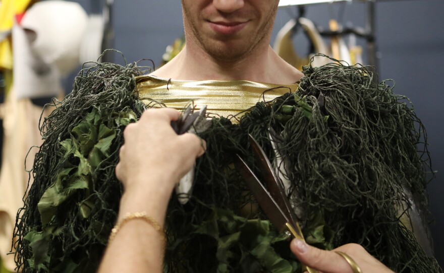 Machine Dazzle removes excess pieces of a costume of Apollo disguised as a mortal – covered in synthetic algae, fishing net and fish – to try to seduce the opera's titular nymph Io.