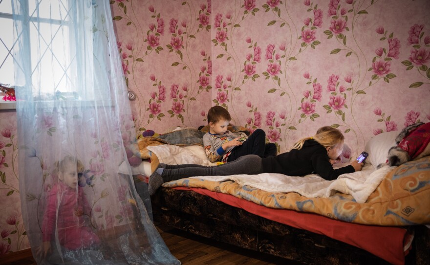 Miroslava Grinik, 8, and her younger brother Kirill, 5, on their smart phones at their new home in Poltava region, which they moved into in October 2022. Their cousin Alisa, plays on the floor by the bed. December 2022.