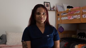 Emely Coleman sits on a bed wearing blue nurse's scrubs.