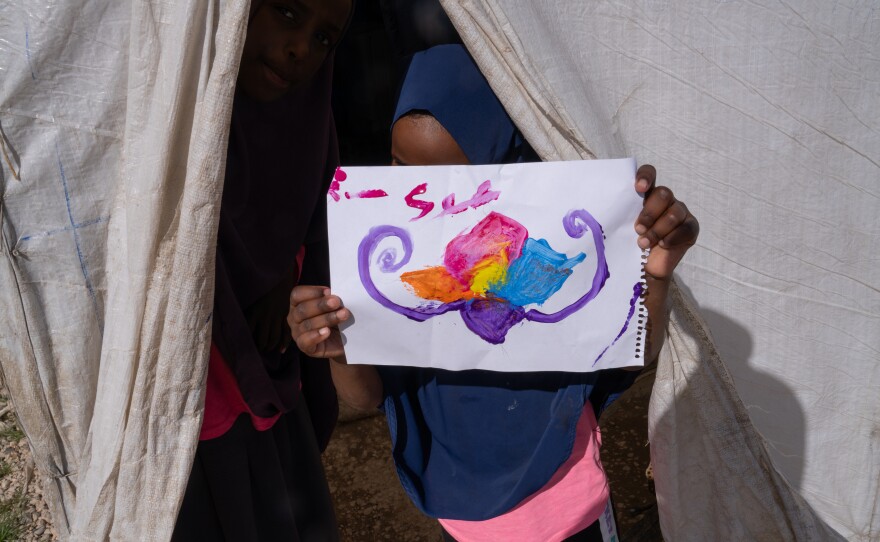 A girl at Roj holds a picture of a flower that she painted. On the back, it says, "hello friends."