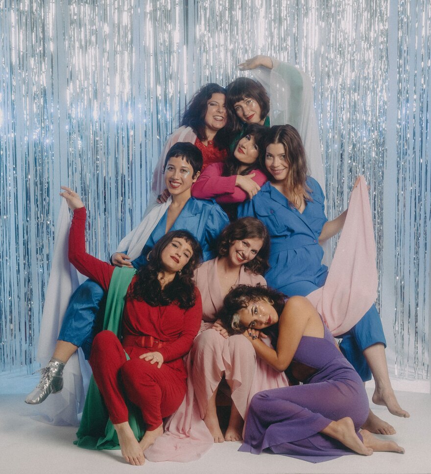 The members of Baby Bushka are dressed in colorful pantsuits in reds, pinks, blues and purples. They're seated in front of a silver-streamered backdrop.