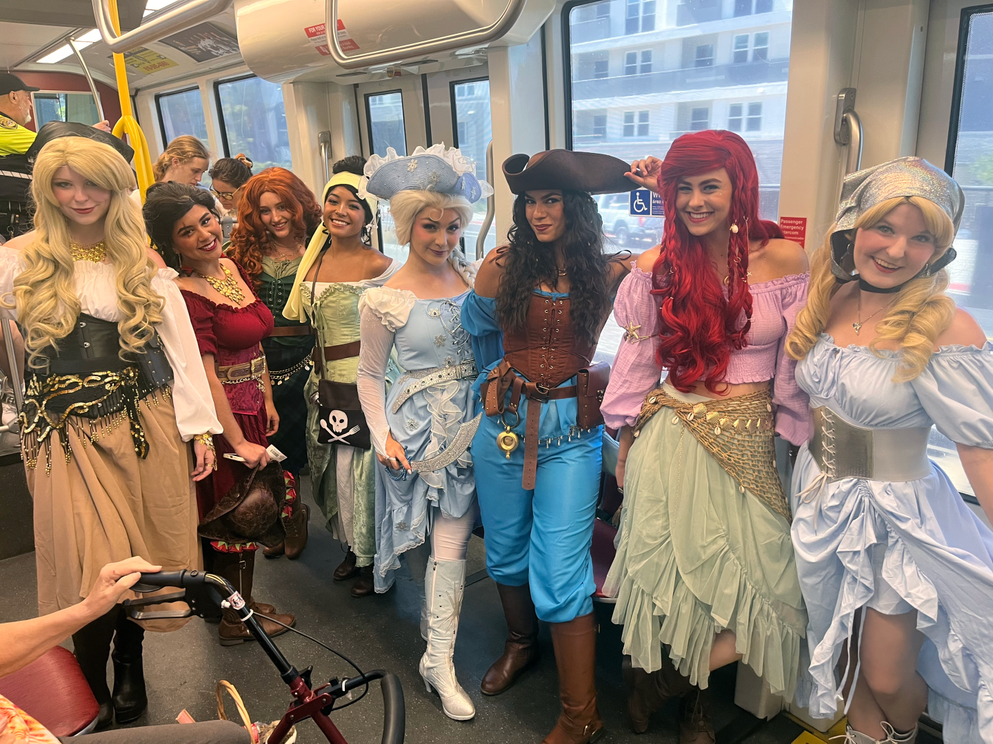 Friends and co-workers ride the Green Line trolley to Comic-Con dressed as pirate-themed Disney princesses on July 24, 2025. From left to right: Marla as Aurora, Alexis as Elena of Avalor, Riley as Merida, Iora as Tiana, Shelby as Elsa, Taylor as Jasmine, Abigail as Ariel and Emmy as Cinderella.