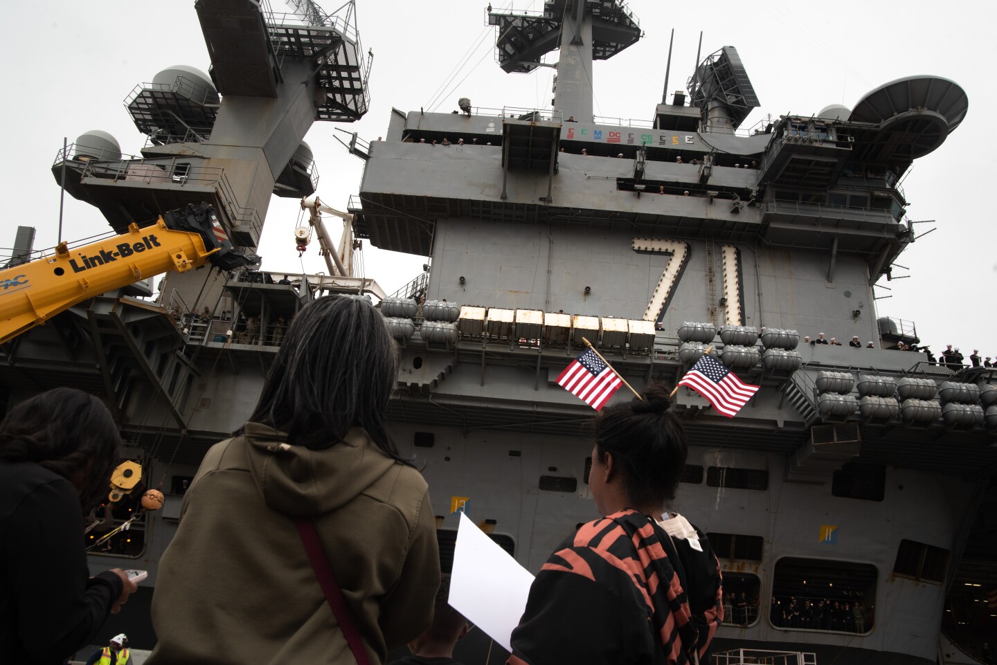 Photo taken from behind three people looking up at the island super structure of the USS Theodore Roosevelt as they wait for their sailor to leave the ship. The woman on the right has two small American flags in her hair.