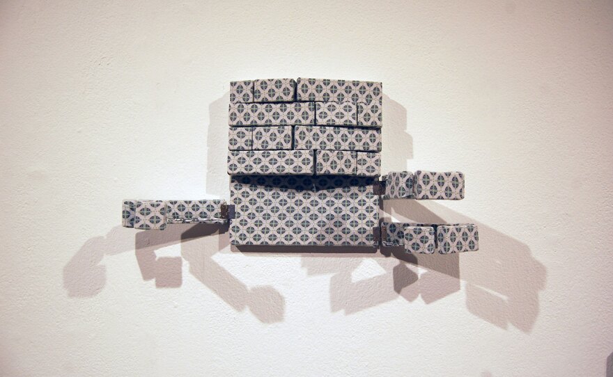 A wall sculpture by Dominic Quagliozzi features a white and navy blue patterned hospital gown stretched across small, hinged wooden frames like a canvas. The pieces are hinged together and movable, to resemble ribs opening up.