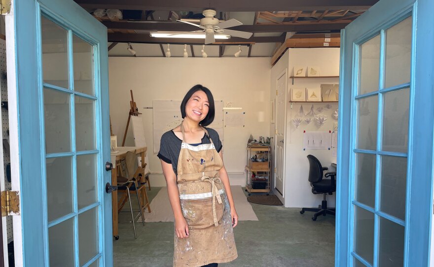 Artist Kaori Fukuyama is shown in her Rolando studio on Aug. 14, 2023. The studio has bright blue French doors, and Kaori stands between them. She has short black hair and wears a paint-splattered, beige-colored apron. Behind her are works in progress and schematic drawings of many of her previous works.