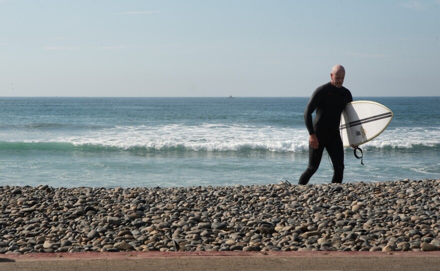 A surfer walks on the rocks between the water and the road named "The Strand" in front of Tyson Street park in Oceanside, California. Coastal erosion has washed away most of the beach's sand. The rocks serve as a replacement until the proposed project REBEACH is underway in late 2026.