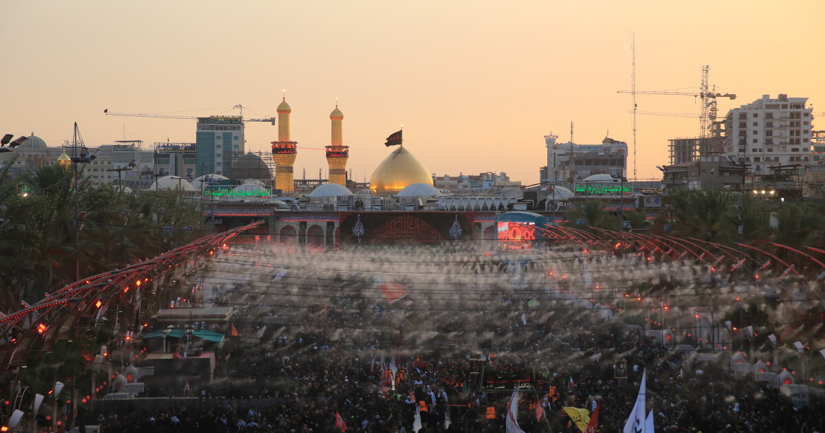Large crowds of pilgrims fill the area between the two holy shrines in Karbala at sunset, with the illuminated dome and