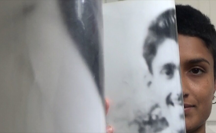 Chloe holds up a photograph of half of her grandfather’s face to her face and looks into the mirror, directly at the camera. There is a striking resemblance between the two of them.