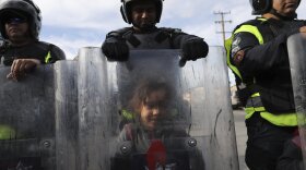 A migrant child plays behind the shield of Mexican police officer in riot gear, after the group she was part of tried to cross the Chaparral border crossing in Tijuana, Mexico, Thursday, Nov. 22, 2018.