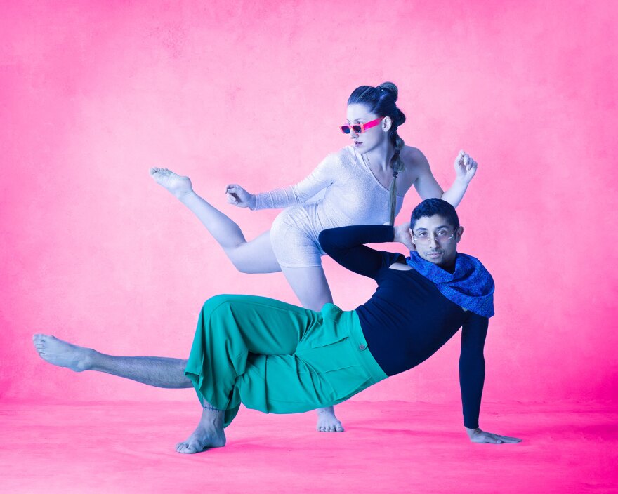 Two dancers wearing sunglasses pose against a hot pink backdrop. One stands on one leg, wearing a pale leotard. One leans on one hand and one foot, wearing green pants, a black leotard and a blue scarf.