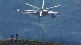 In this photo provided by the Santa Barbara County Fire Department, a Skycrane helicopter passes firefighters atop a hillside while coming in for a water drop below E. Camino Cielo in Santa Barbara, Calif., Tuesday, Dec. 19, 2017.