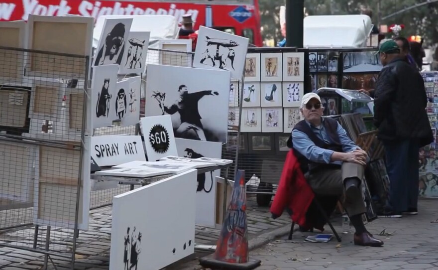 An image from a video posted by Banksy shows a man representing the artist staffing a sidewalk stall featuring signed works for $60. Banksy says he only made $420 Saturday, with one customer negotiating a 2-for-1 discount.