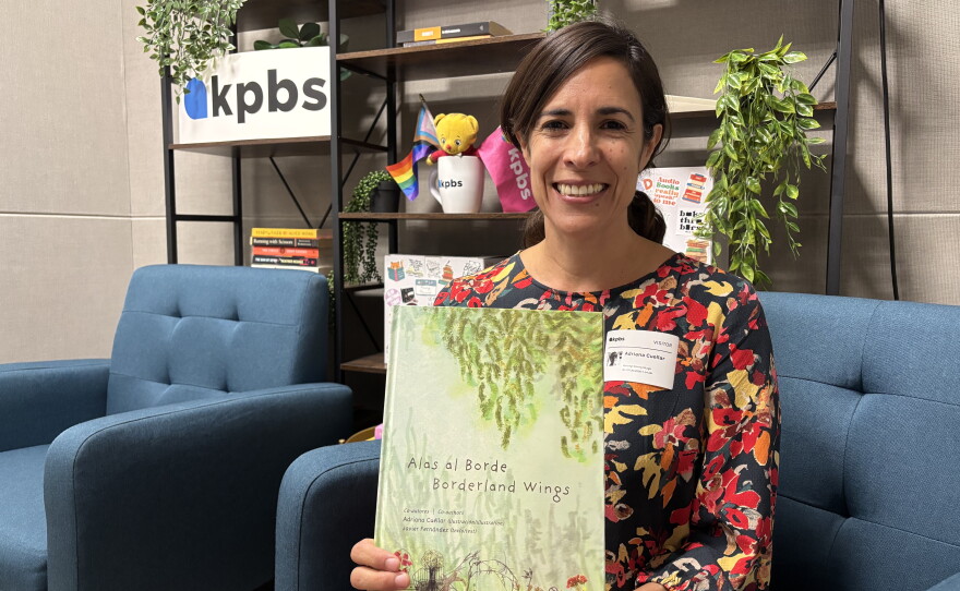 University of San Diego professor and founder of the Border Design Lab, Adriana Cuéllar, holds up her book, "Alas al Borde: Borderland Wings" in the KPBS social media studio, July 30, 2026.