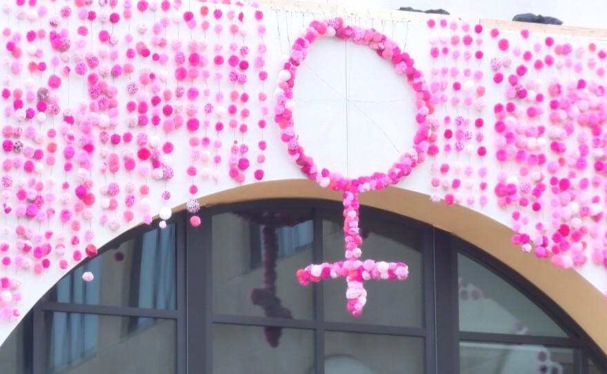 Pink pompoms hang from a building, surrounding pompoms shaped into the woman symbol.