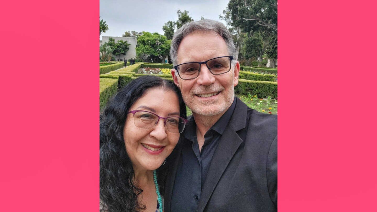 A woman with shoulder length dark hair wearing glasses smiling next to a man wearing glasses and a black shirt and suit coat. The two are standing in front of greenery at Balboa Park.