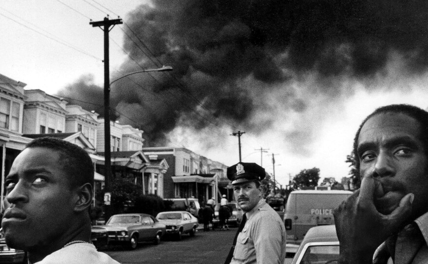 The scene at the corner of 62nd and Larchwood, in the afternoon following the dropping of bomb on MOVE headquarters, May 13, 1985. Smoke pours out over 6200 block of Osage Avenue as fire begins following police helicopter dropping a bomb onto roof of the MOVE house.