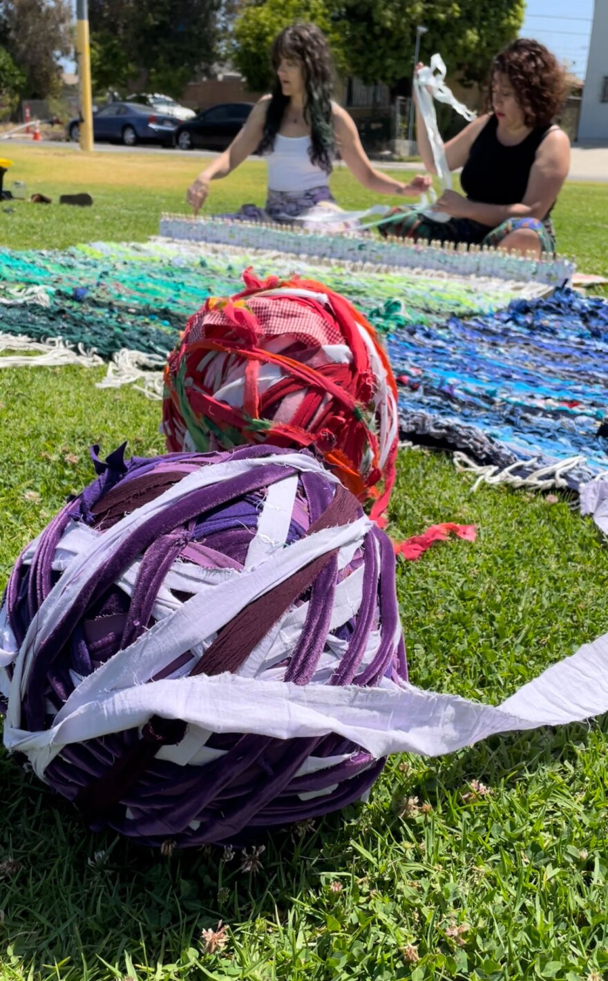 large balls of "yarn" made from strips of clothing with two artists, seated and weaving at a loom.