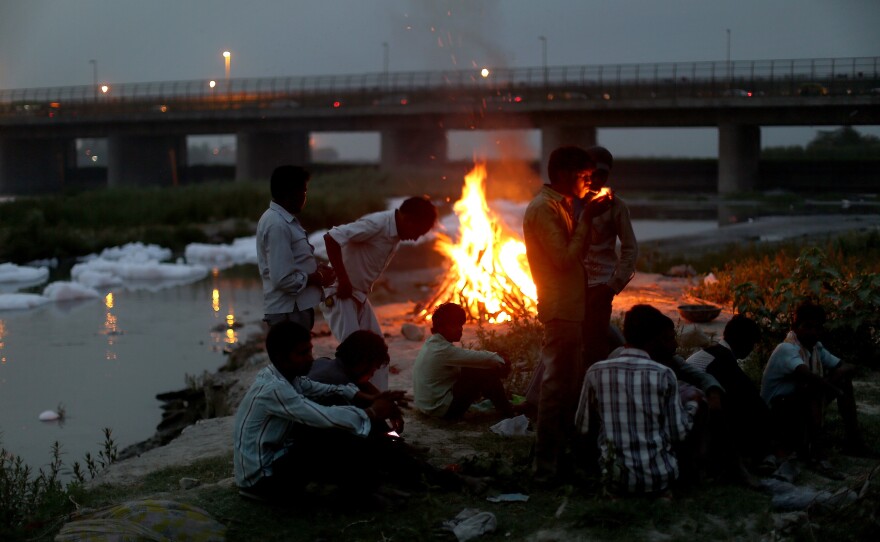 Men watch the fires of a cremation along the banks of the Yamuna River against the backdrop of the Wazirabad Barrage and floating industrial waste.