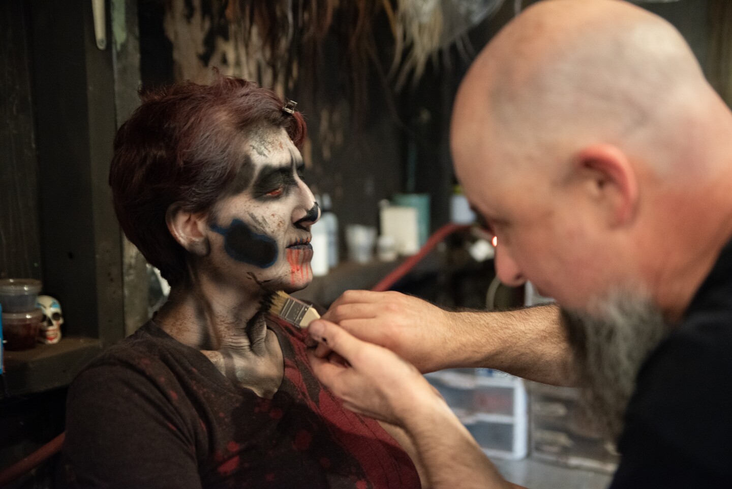 Makeup artist Stuart Hirsch applies some deathly texture with a brush to the airbrushed graveyard makeup for KPBS reporter Beth Accomando at The Haunted Trail in Balboa Park on Sept. 25, 2025.