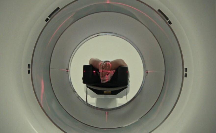 The camera looks down the several mechanical and concentric circles of a large MRI machine to a lone figure in the middle. The figure lies on their back, everything obscured except their hands, which press together towards the camera, above the figure's head. Red lights create a bull's eye on the figure's hands, giving the entire image a science-fiction feel.