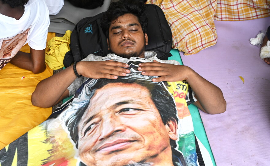 A reclining protestor covers himself with a banner printed with a portrait of Sonam Wangchuk, a prominent activist who was forcefully transferred to a hospital by the police as he staged a hunger strike in support of a movement led by Cockroach Janta Party, at a protest in New Delhi, India, Wednesday, July 22, 2026.