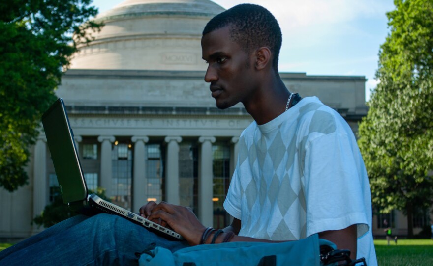 Billy, a young Black man from Rwanda, works on his laptop outdoors on a lawn at MIT (the Killian Court lawn). The gray dome-shaped building with Greek pillars fills the background. He has short, curly, black hair and is wearing a checkered white and gray t-shirt.