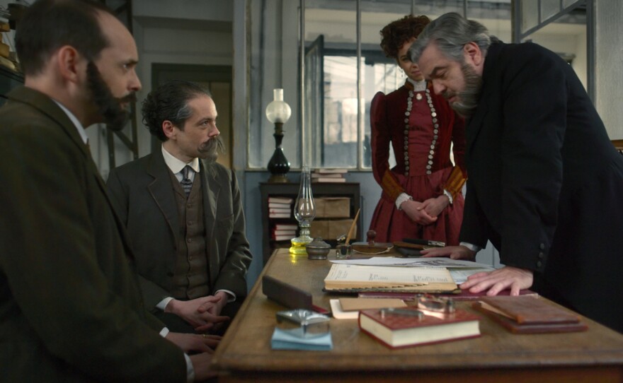 Gustave Eiffel is behind his desk, his daughter on his right, his associates are showing him calculation for the tower.