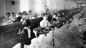 Women sitting at sewing machines in the Triangle Shirtwaist Factory, with piles of white fabric in front of them. A sign attached to the ceiling at the back says "Fire Escape" and has a hand pointing to a window.