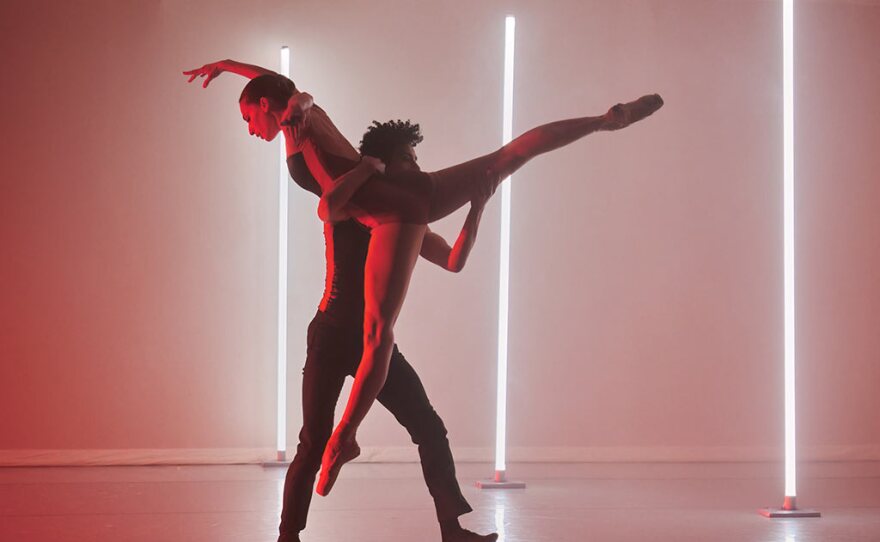 Dancers from The Rosin Box Project are shown in an undated photo. One dancer is lifting another, who is wearing pointe shoes and has both legs extended in the splits in the air. Her hand is curved up above her head. Three columns of bright white light are positioned on the stage behind the dancers.