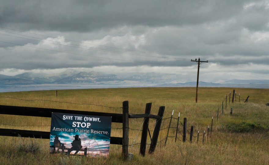 A "Save the Cowboy" sign is posted along a fence. The "Little Rockies" on the Fort Belknap Indian Reservation are seen in the distance.
