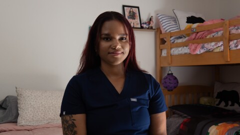Emely Coleman sits on a bed wearing blue nurse's scrubs.