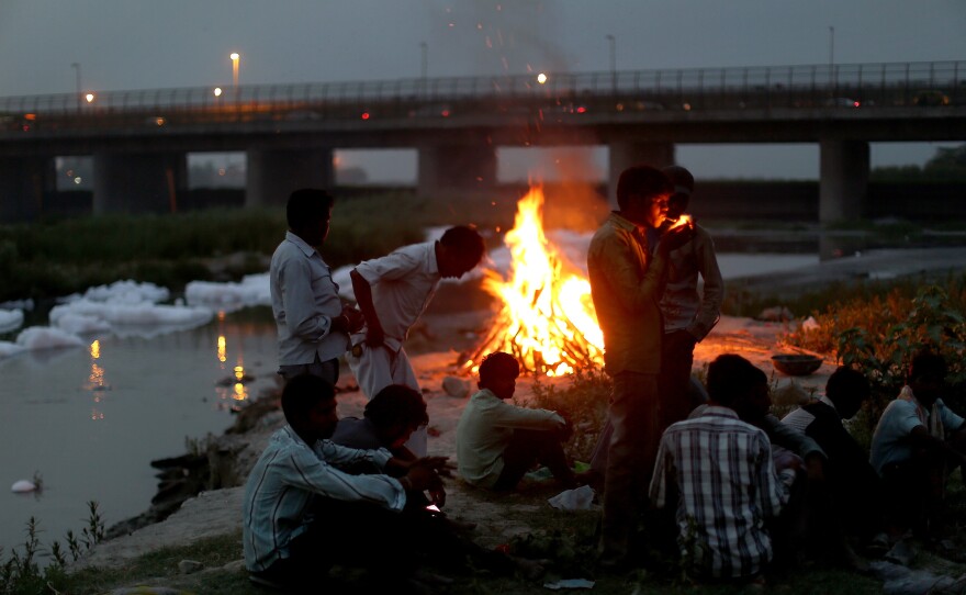 Men watch the fires of a cremation along the banks of the Yamuna River against the backdrop of the Wazirabad Barrage and floating industrial waste.