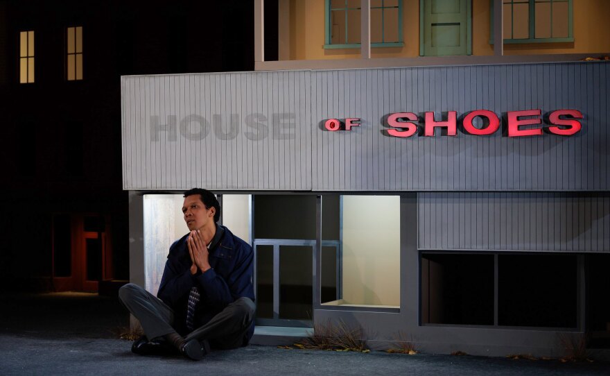 A man is seated, cross-legged, on the stage. The set is a miniature-scaled run-down shoe store, "House of Shoes," and the sign is partially lit. Tufts of weeds grow at the base of the building. The man holds his hands together in prayer position, deep in thought.