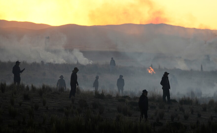 Anti-government protesters standing in a field are silhouetted against clouds of tear gas launched by police, near El Alto, Bolivia, Saturday, May 16, 2026.