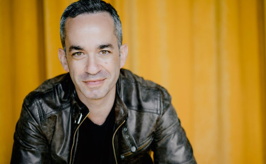 Inon Barnatan is shown against a yellow backdrop, wearing a black jacket and shirt. He is looking at the camera and smiling, and has black/graying hair and light skin.