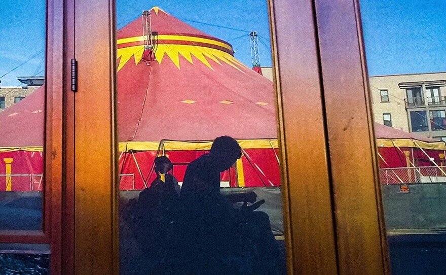 In a reflection of an unmarked storefront is a grayish silhouette of a man using an electric wheelchair. Behind the man is a spectacular red and yellow circus tent.