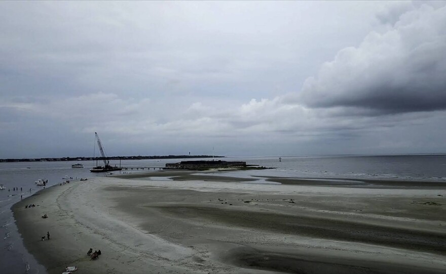 A long shot of a sandy shore with people scattered along it, and in the far distance, the remains of Fort Sumter can be seen. A small boat is anchored in the water with a crane overhead. The sky is overcast with clouds, and a rocky outcropping juts into the water, creating a dramatic contrast between the land and sea.