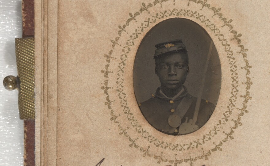 A black-and-white photograph of John Walls. He is wearing a buttoned-up jacket with epaulets on the shoulders and a kepi. His kepi is on the right side of his head. He has a shoulder strap on his left side. His name is inscribed below the photograph on the same page.