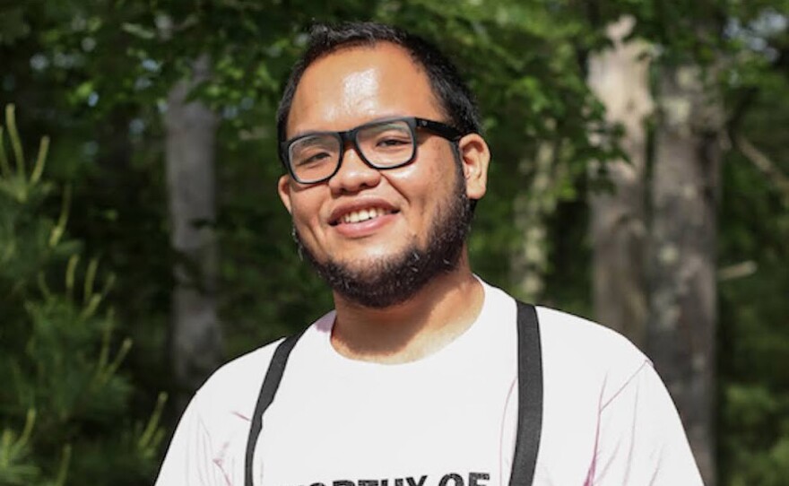 A male-presenting person with olive complexion, black hair and beard stands in front of trees. They are wearing glasses, suspenders, and a pink shirt with the text “Worthy of Humanity.” Descripción: Una persona de aspecto masculino y tez aceitunada, con pelo y barba negra, posa frente unos árboles. Lleva gafas, tirantes y una camisa rosa que lee "Digno de la Humanidad".
