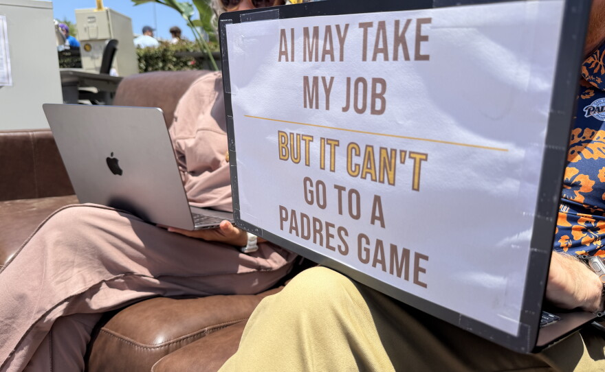 Remote workers at the San Diego Padres game on Wednesday, June 10 have a sign on a laptop reading "AI may take my job but it can't go to a Padres game."