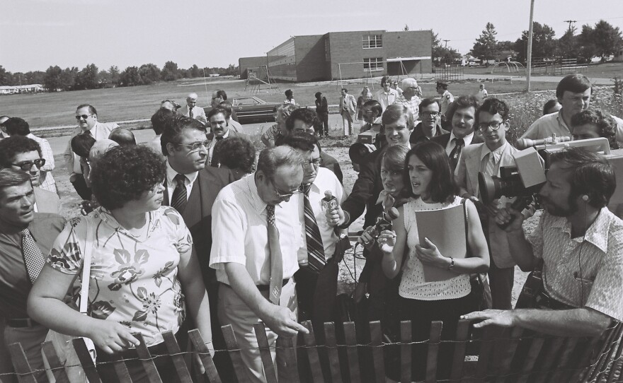 Lois Gibbs (holding notebook), standing with a group of federal officials and politicians, speaks to the media with the 99th Street School in the background. Debbie Cerrillo Curry is standing against the fence on the left. August 1978.