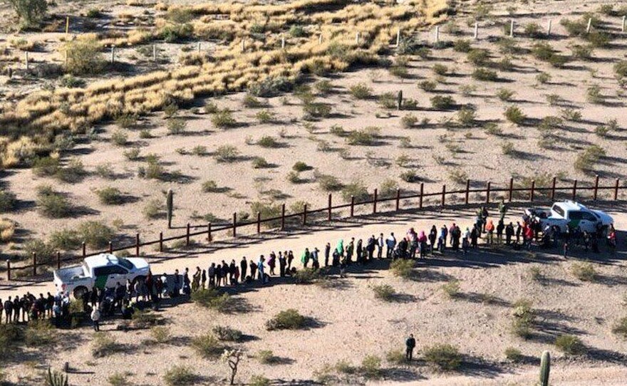 Migrants line up after being apprehended along the U.S.-Mexico border near Lukeville, Ariz. Mexico is at the top of the image, beyond the border fence.
