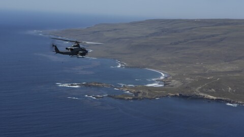 A Marine Corps Ah-1Z SuperCobra attack helicopter flies from the left toward the western rocky shore of San Clemente Island.
