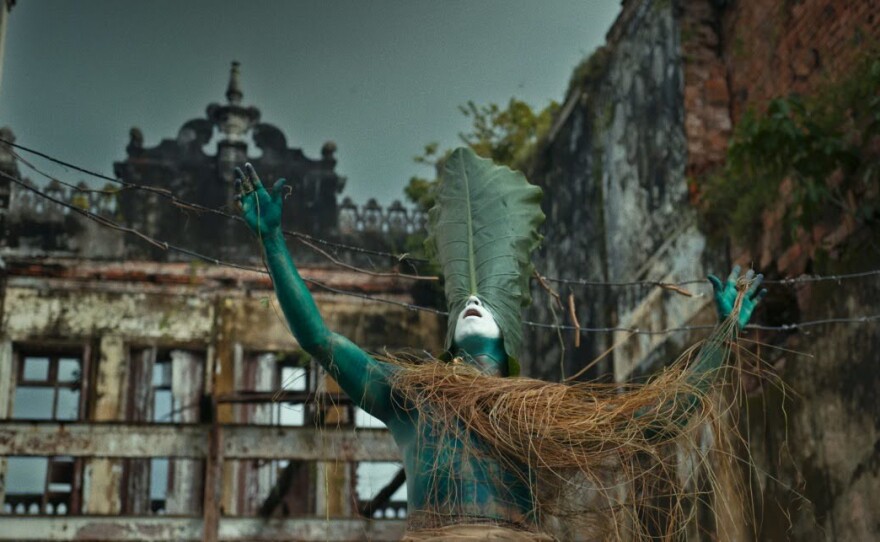 Surrounded by crumbling buildings of brick, concrete, windows and green ivy, a figure strikes a pose with arms held up, palms visible and faces the viewer. Their body is painted green and strewn with long dried grass that covers the left arm and part of the chest. Their face, partially obstructed by a large green leaf drawn over the eyes, is painted white; only the nose, mouth and chin are visible. The blue sky looms overhead, calling attention to the dramatic architectural pointed feature sitting atop the center of the building.